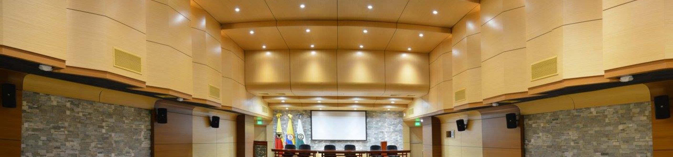 Auditorio de la Universidad La Gran Colombia con escenario, mesa y pantalla de proyección; butacas verdes para congresos y conferencias.